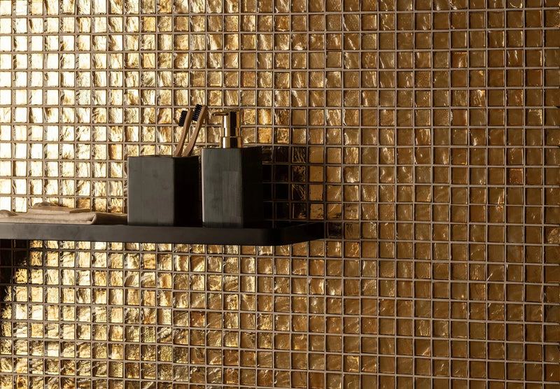 Black rectangular soap dispenser with metallic gold pump on dark shelf against shimmering gold mosaic tiles background