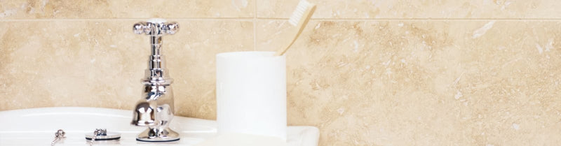 Chrome-finished faucet with cross-shaped handle beside white cylindrical toothbrush holder in travertine tiles setting