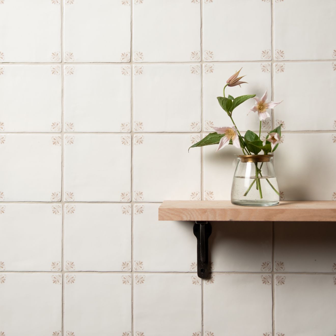 Glass vase with flowers on a wooden shelf against a tiled wall.