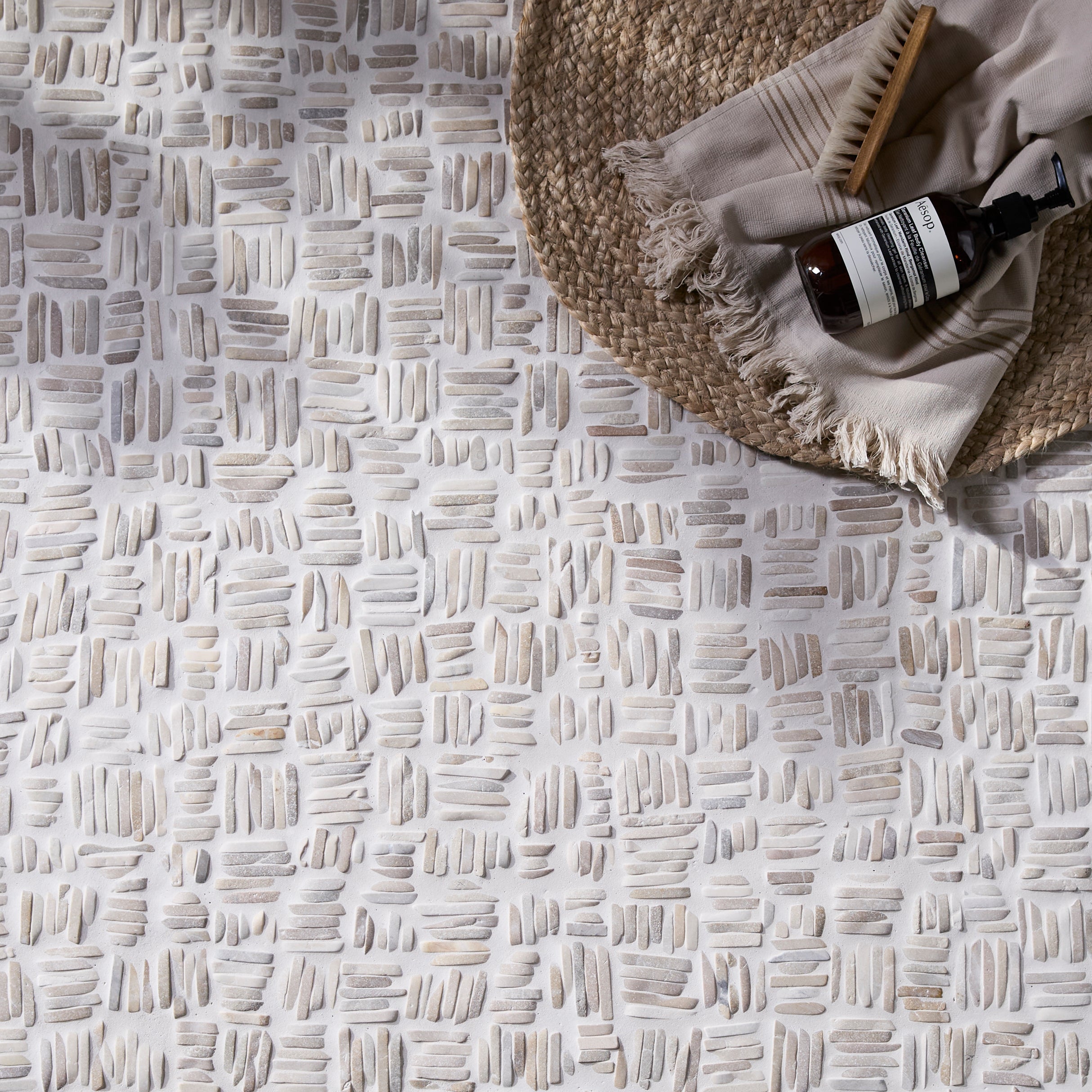 Textured floor in Enso Organic Basketweave Limestone Mosaic Tiles with a woven basket and bottle on a light gray background