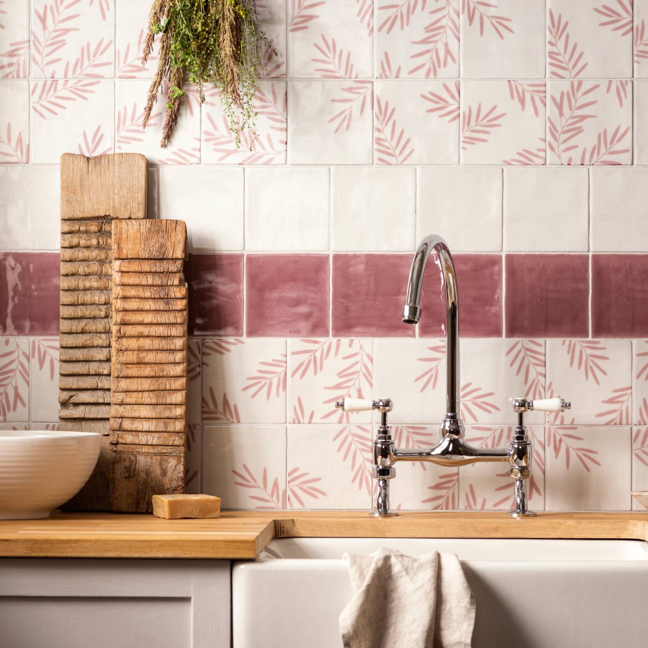 Kitchen sink area with wooden cutting board, soap, and towel against a tiled wall with pink and white pattern.