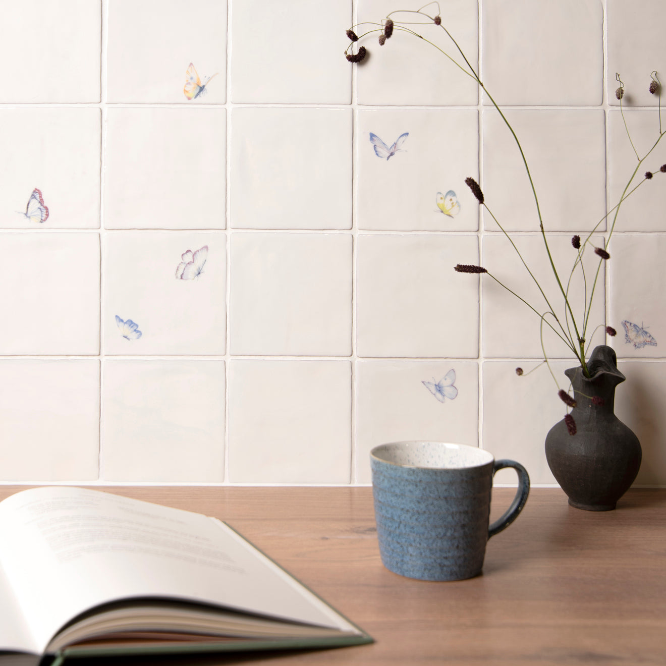 Ceramic tiles with butterfly designs on a wall above a wooden surface with a mug and vase.