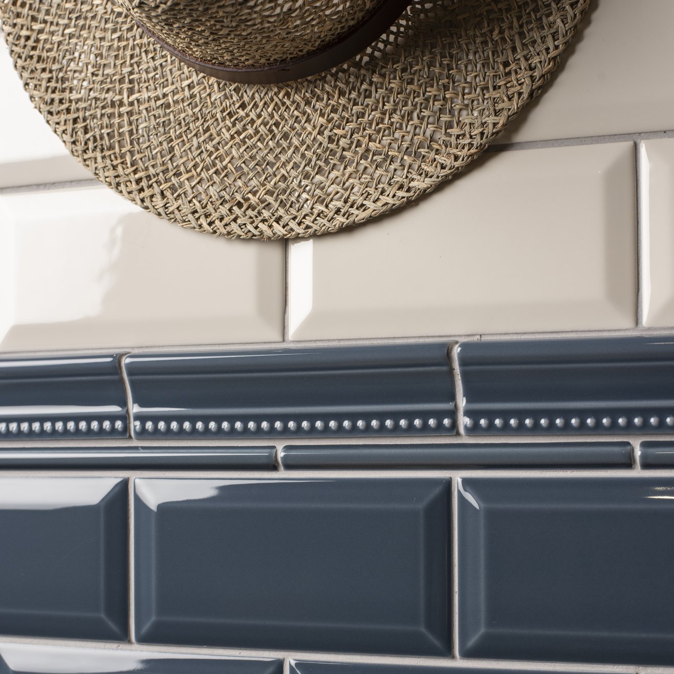 A close-up of glossy rectangular ceramic subway tiles in two colors: a dark slate blue lower section and a warm cream upper section, separated by a decorative white beaded tile band. A woven straw hat is partially visible at the top edge.