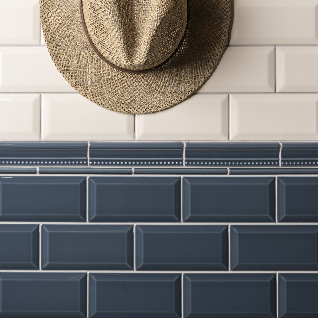 Two styles of subway tiles shown in a split layout: white glossy rectangular tiles on the top half with a woven straw hat hanging on the wall, and dark blue-gray glossy rectangular tiles on the bottom half.