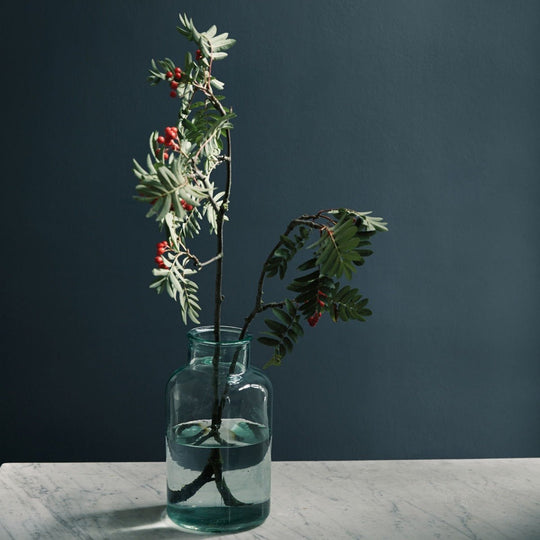 A clear glass vase named carbon blue with a rounded body and narrow neck holding green branches with red berries