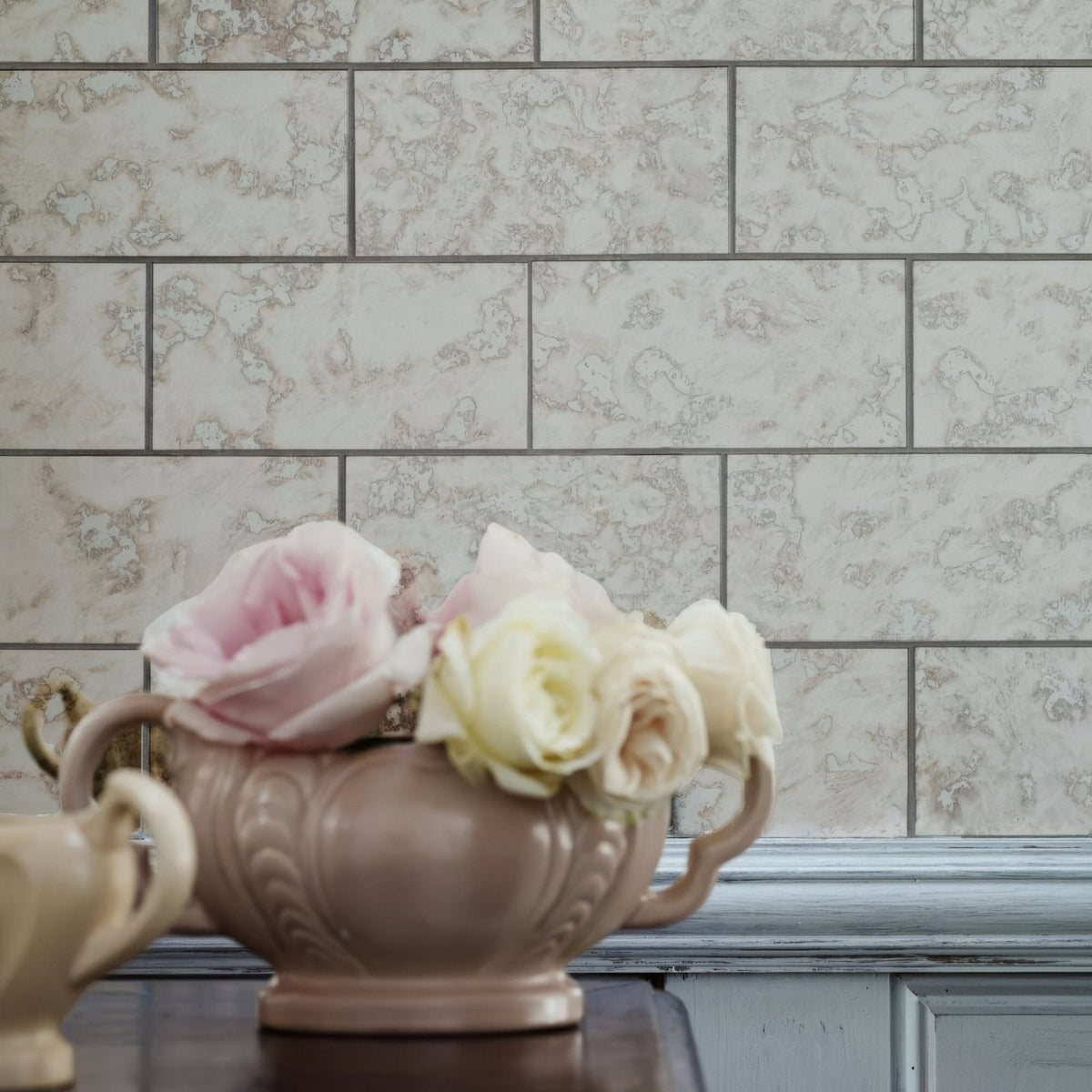 A beige ceramic vase with ornate embossed detailing and handles holding pink and cream roses in deco glass pewter tile