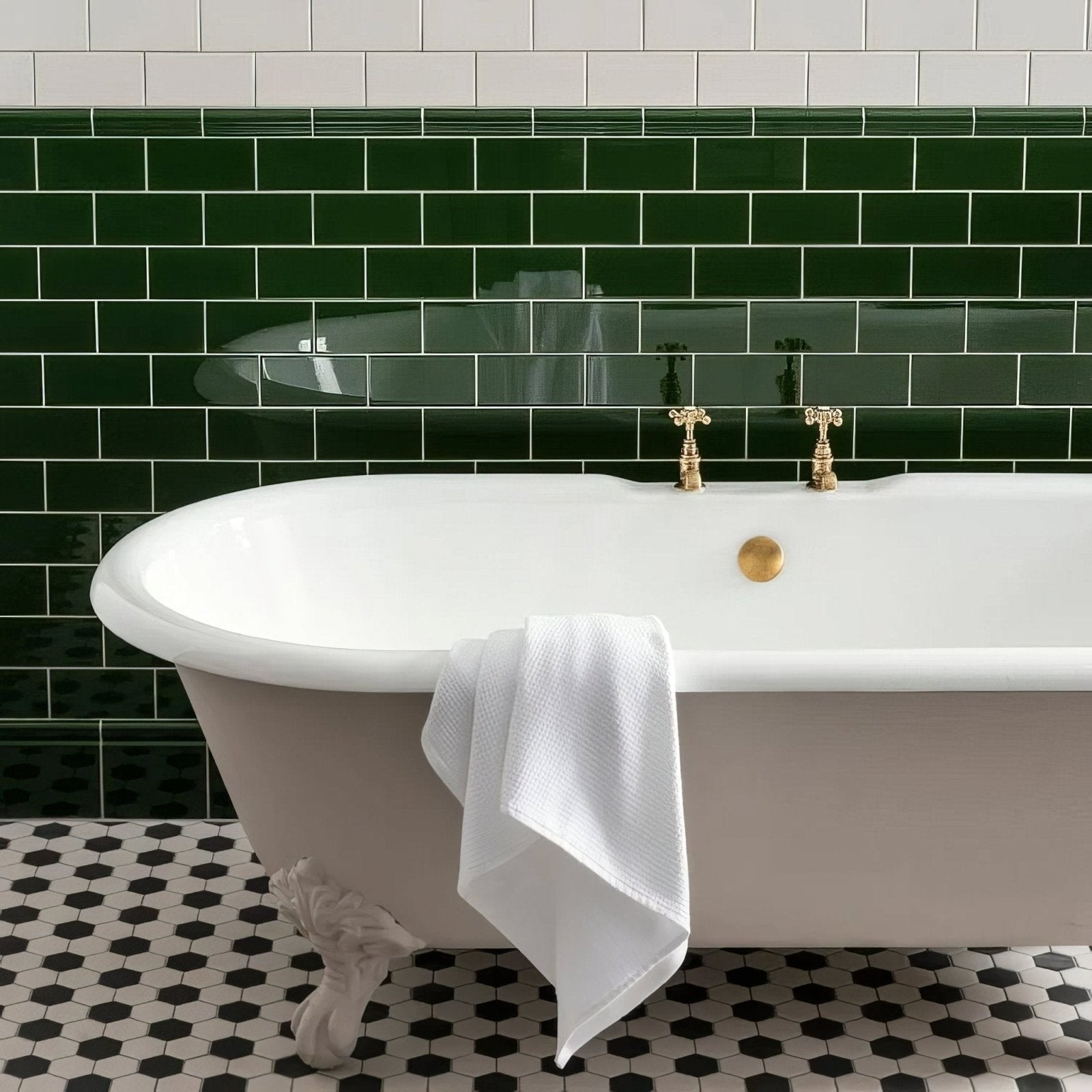 A classic white clawfoot bathtub with gold fixtures and a plush white towel in an Edwardian green half tile setting