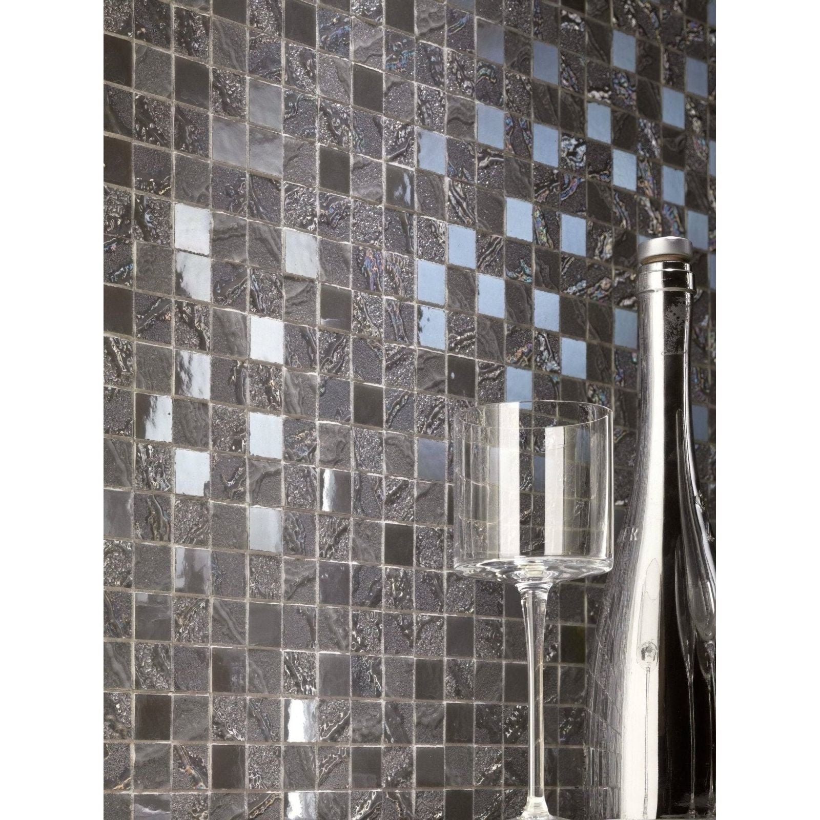 A sleek transparent glass wine bottle with a metallic cap next to an elegant stemmed wineglass on dark mosaic tiles