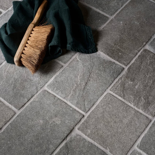 A rustic wooden-handled broom with natural brown bristles rests on dark green cloth over Galway Limestone Cobble seasoned finish tiles