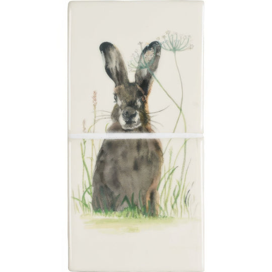 A rectangular ceramic tile panel titled hare in hiding 2 featuring a watercolor brown hare with wildflowers and grass