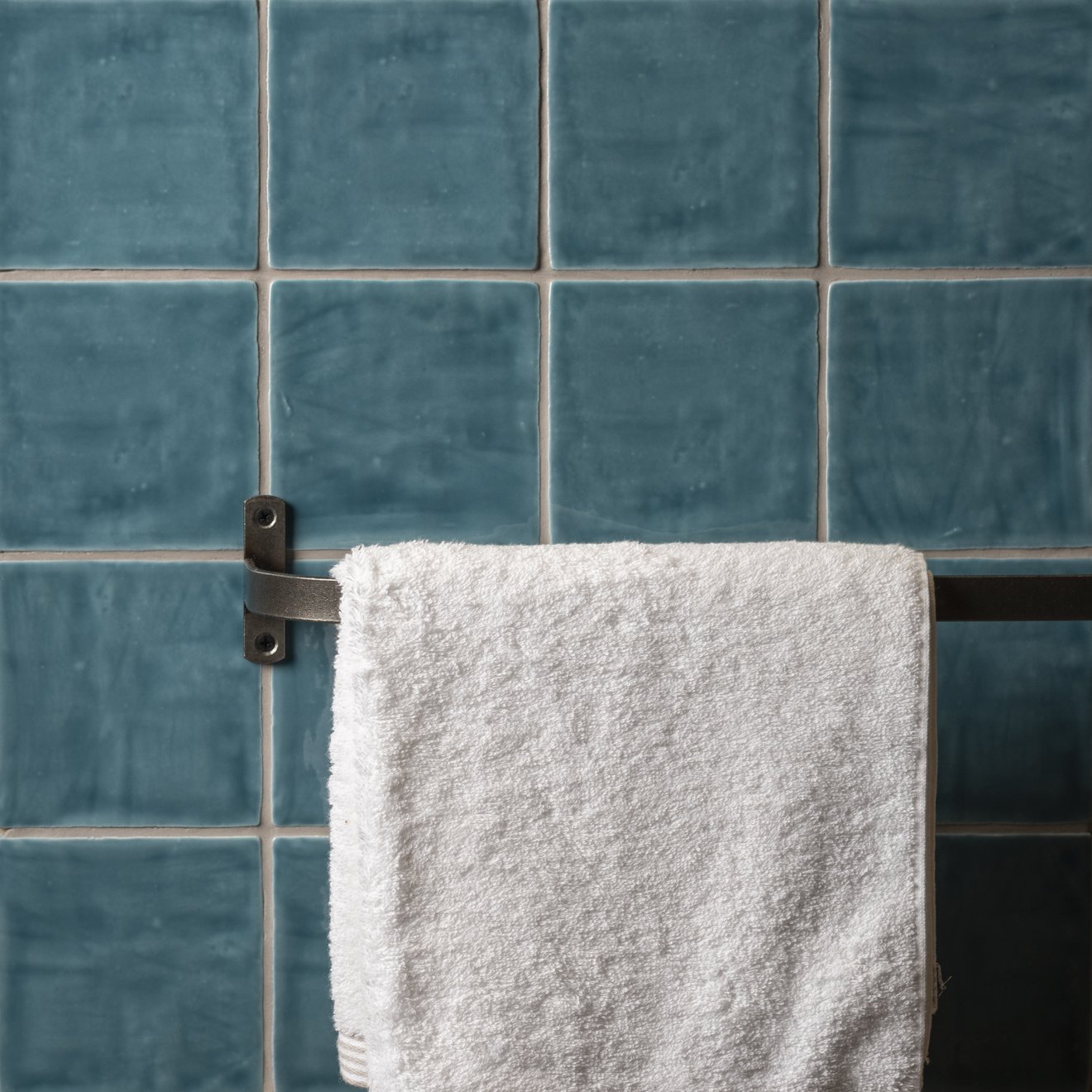 A plush white towel hangs on a dark metal rack against textured teal lagoon field tile wall