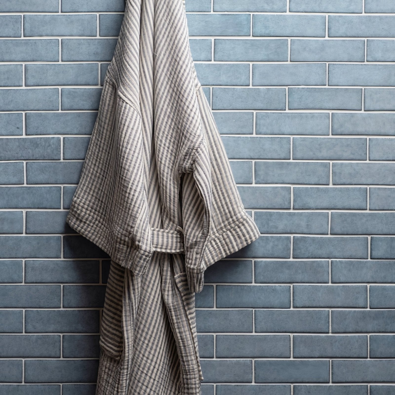 A soft beige and gray striped cotton robe with matching belt hangs against a blue tiled wall in livid brickbond mosaic