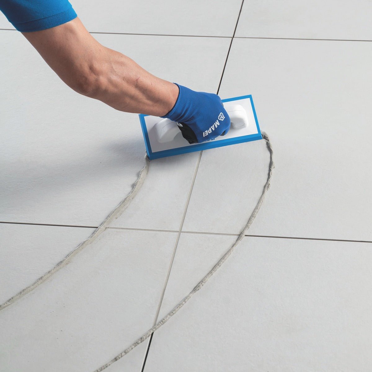 Blue-handled tile squeegee with white rubber blade smoothing grout on light gray tiles using Mapei Ultracolour Plus Grouts