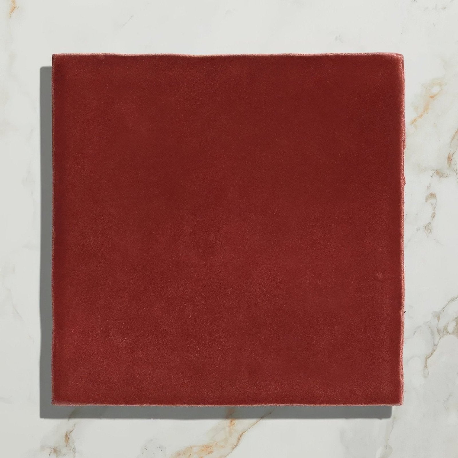 A square matte-finished terracotta ceramic tile with uneven edges from maroc ceramic bordeaux on a marble surface