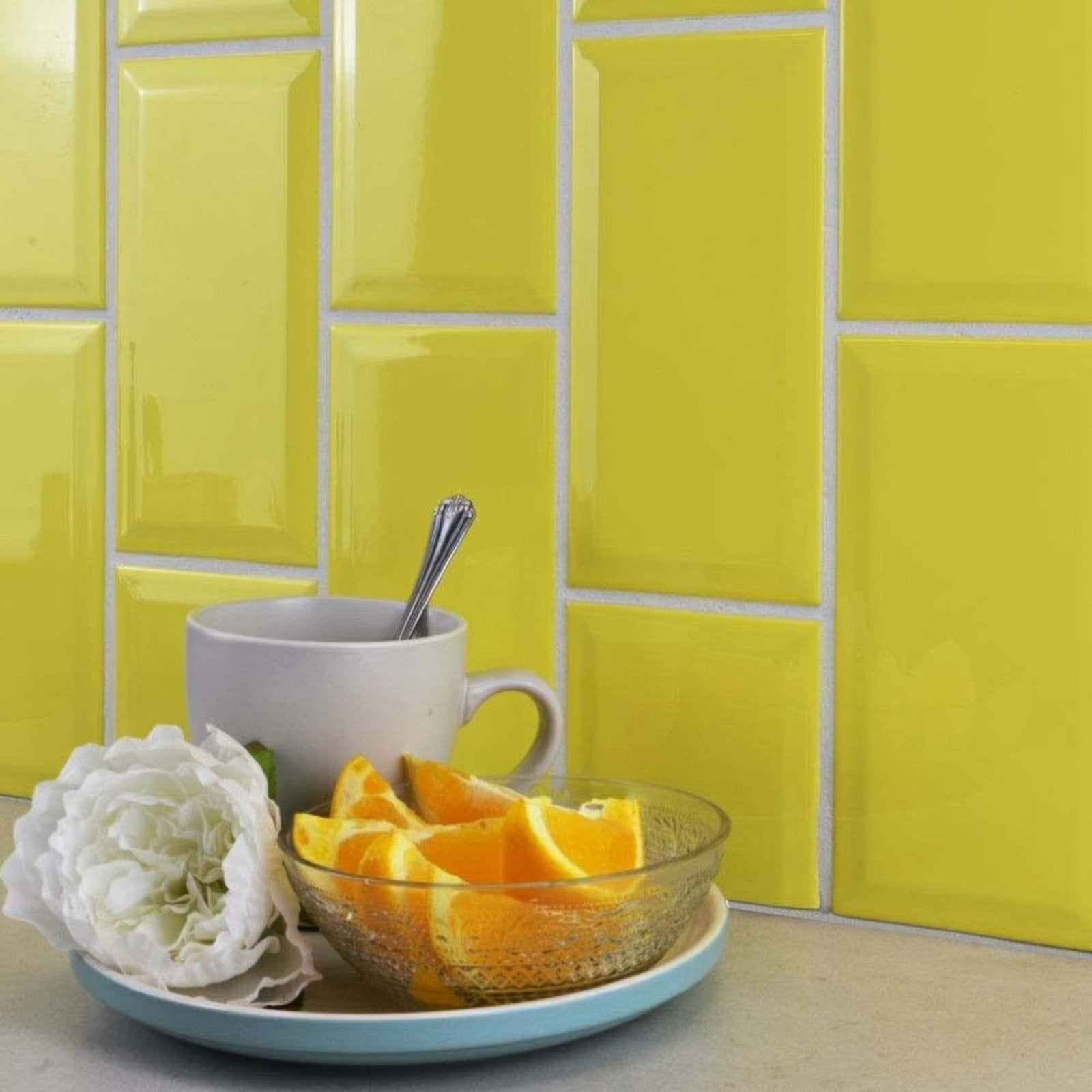 A clear glass bowl with bright orange slices on a light blue plate next to a white flower and gray mug metro limon gloss 100x200