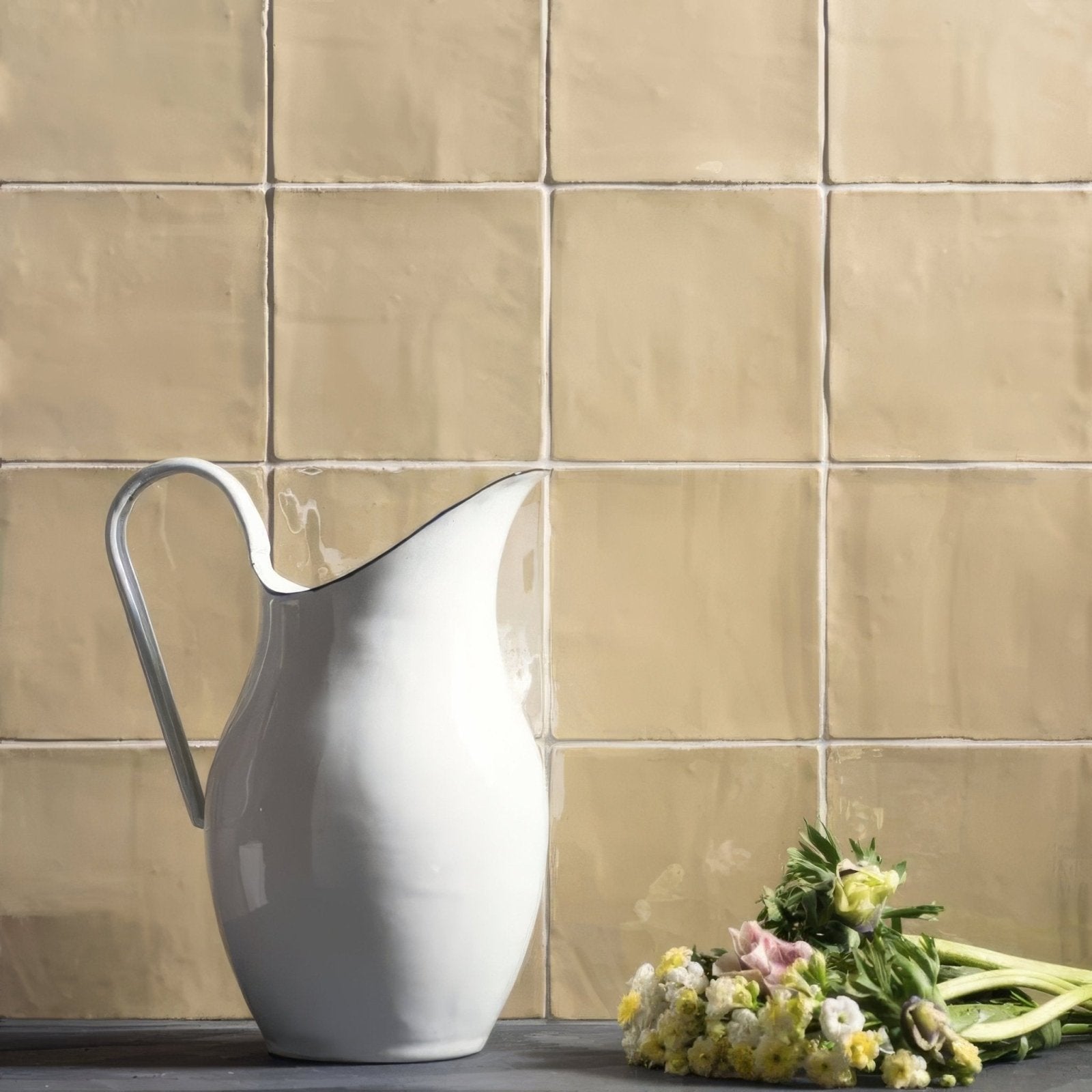 A sleek white ceramic pitcher with a curved metallic handle stands against warm beige mimosa field tile backdrop