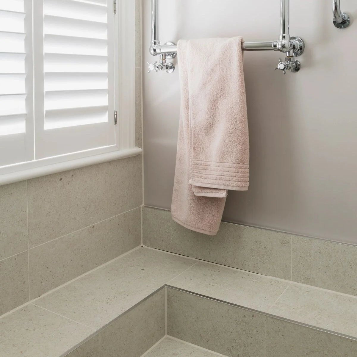 A soft blush pink Piazza Bone towel with plush texture and folded hem hangs from a chrome towel rack