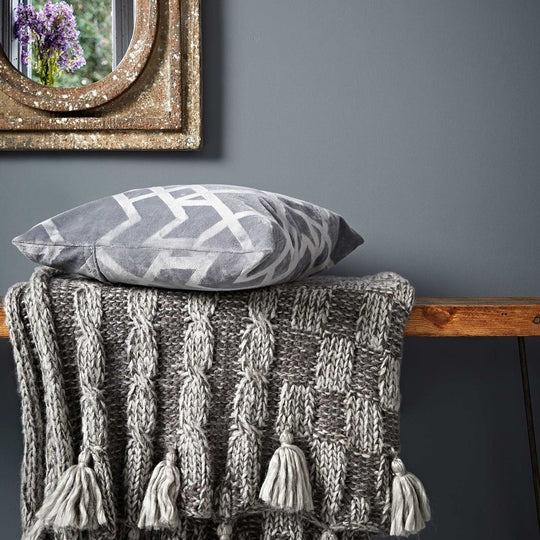Gray plumbago cable knit throw blanket with matching tassels draped over wooden surface
