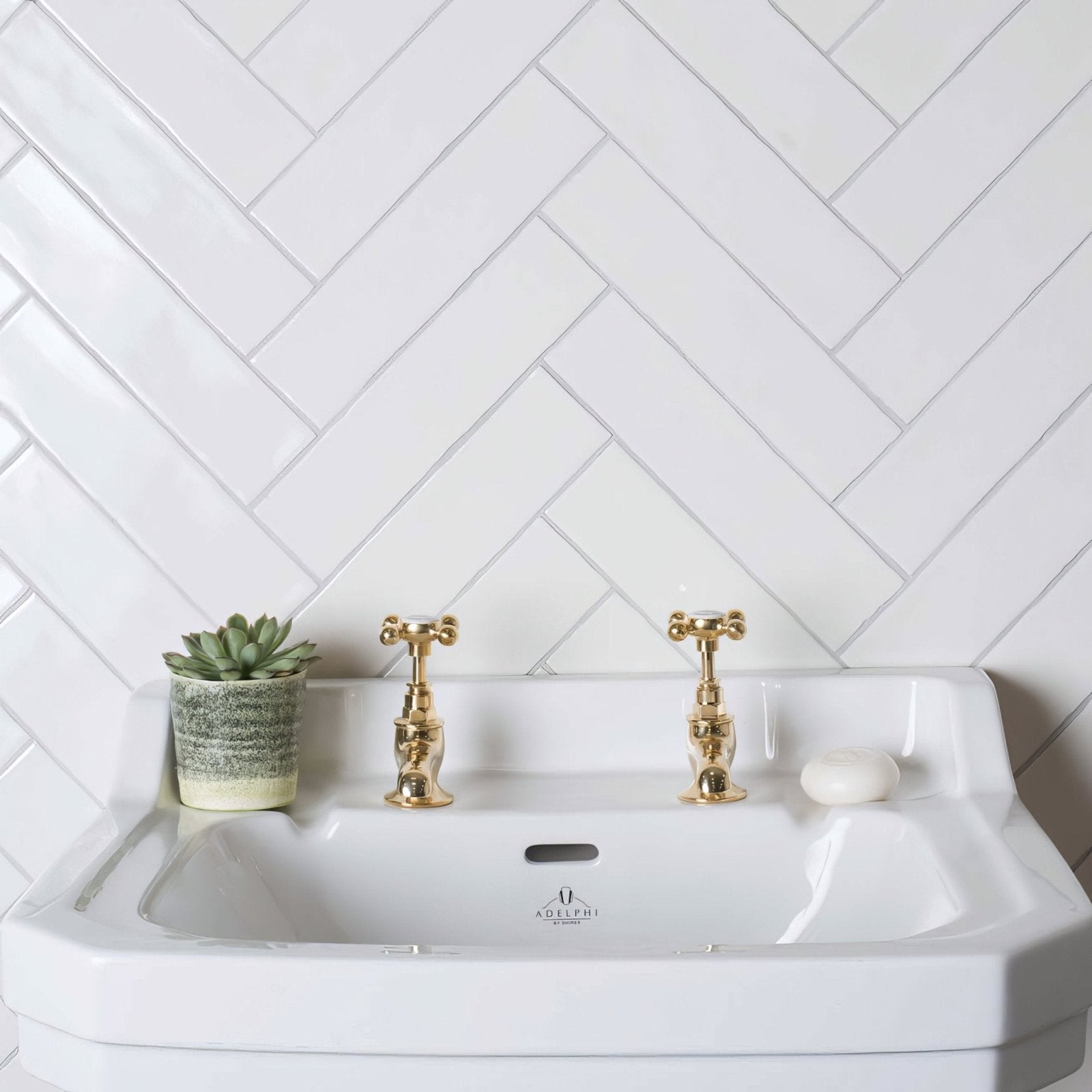 White ceramic sink with gold-finished cross-handle faucets and subtle adelfri logo paired with large brick tile porcelain