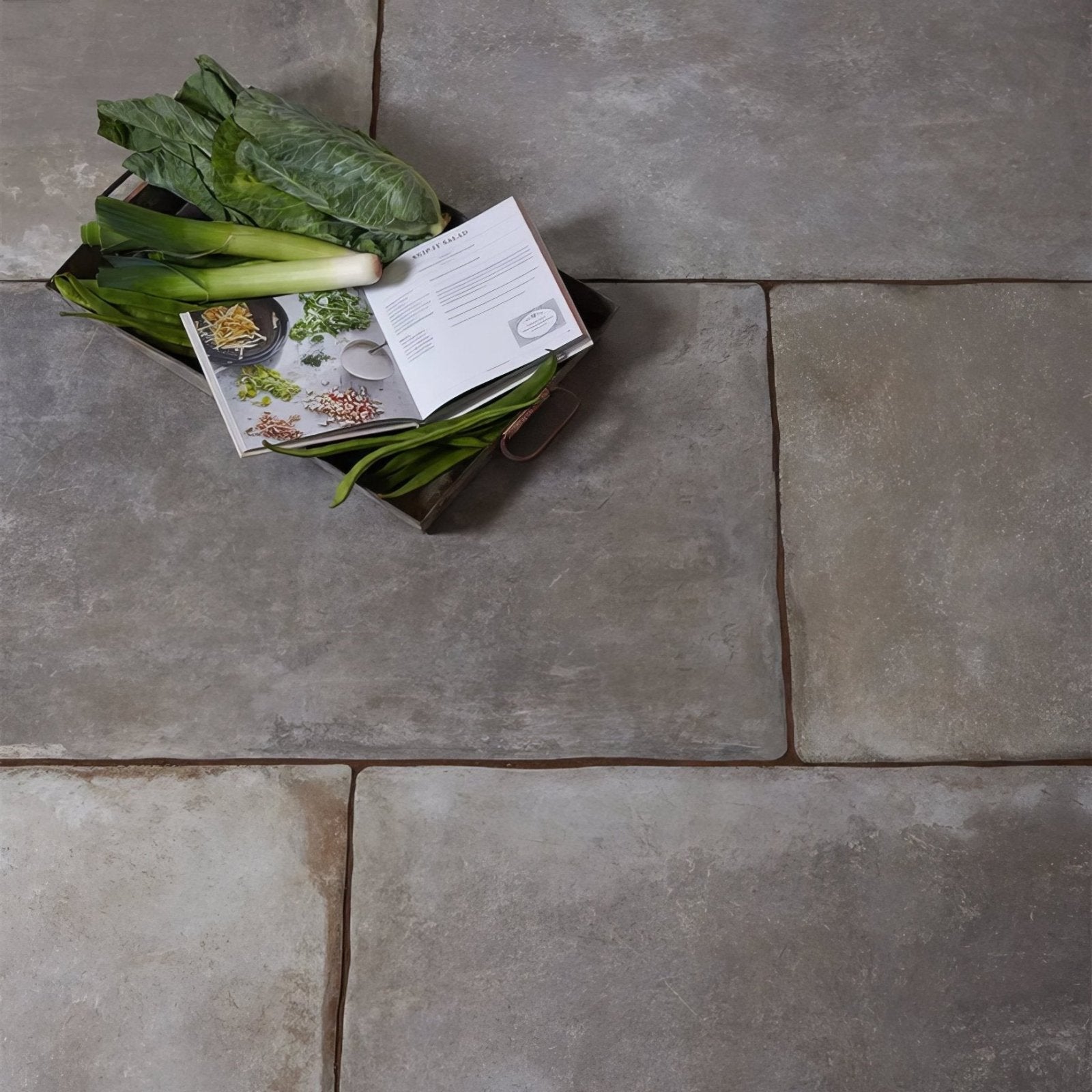 A rustic metal tray with fresh green vegetables and an open cookbook on a provence paving porcelain cenere surface