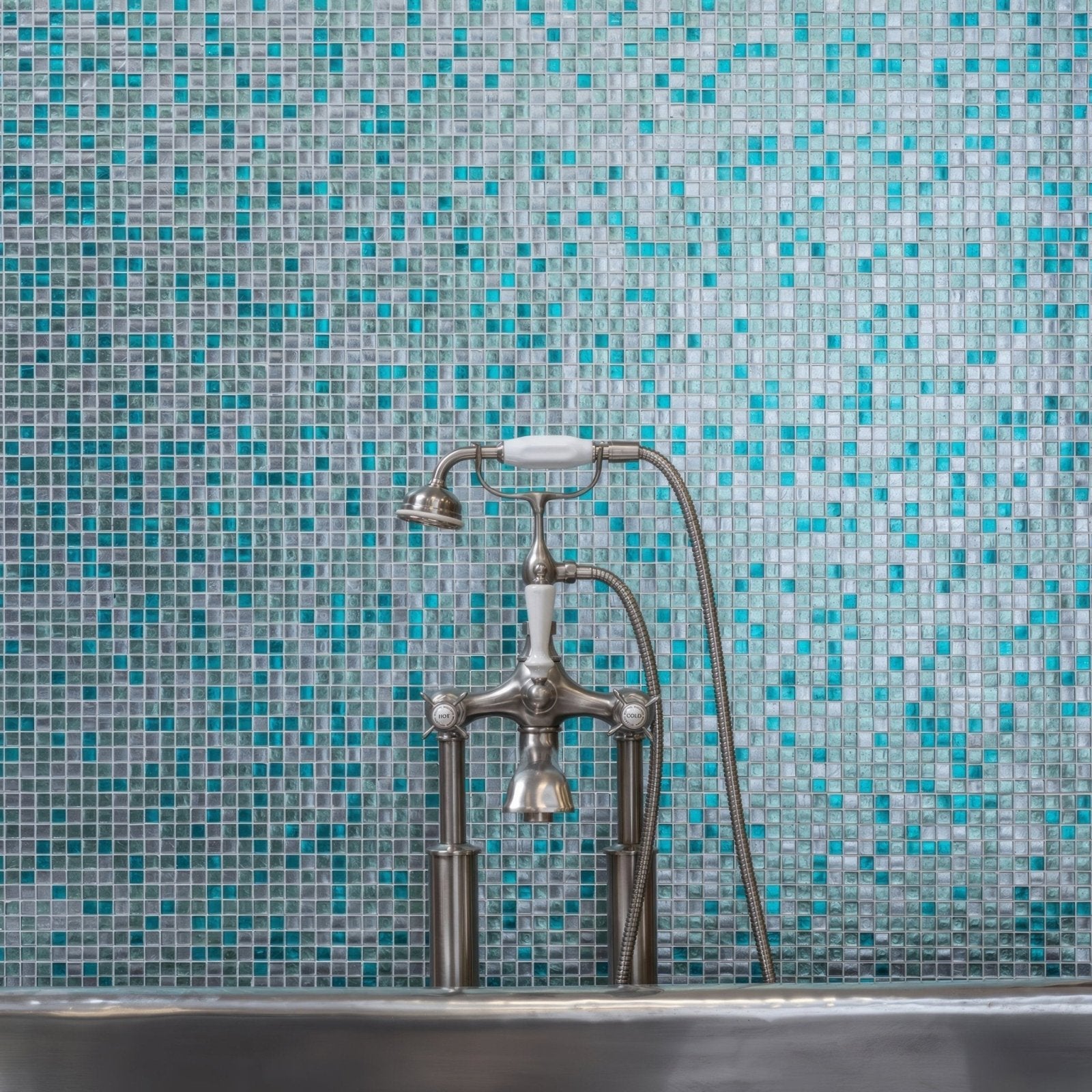 Vintage-style chrome faucet with white ceramic handles and flexible hose against turquoise ripple mosaic tiles