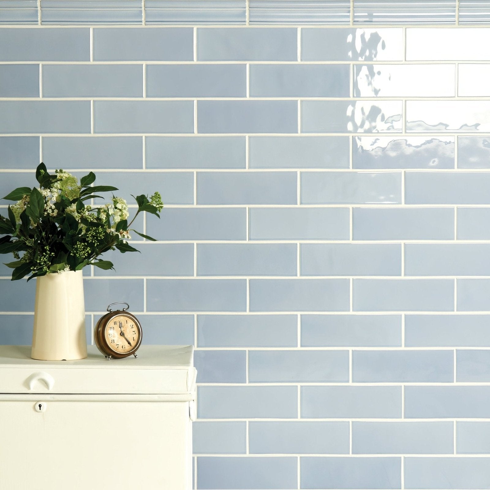 A cream-colored ceramic vase with green foliage and small white flowers in the brick tile snape style