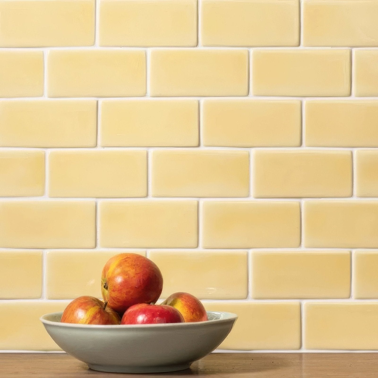 A light gray ceramic bowl filled with red and yellow apples on a wooden surface beside soft yellow half tile