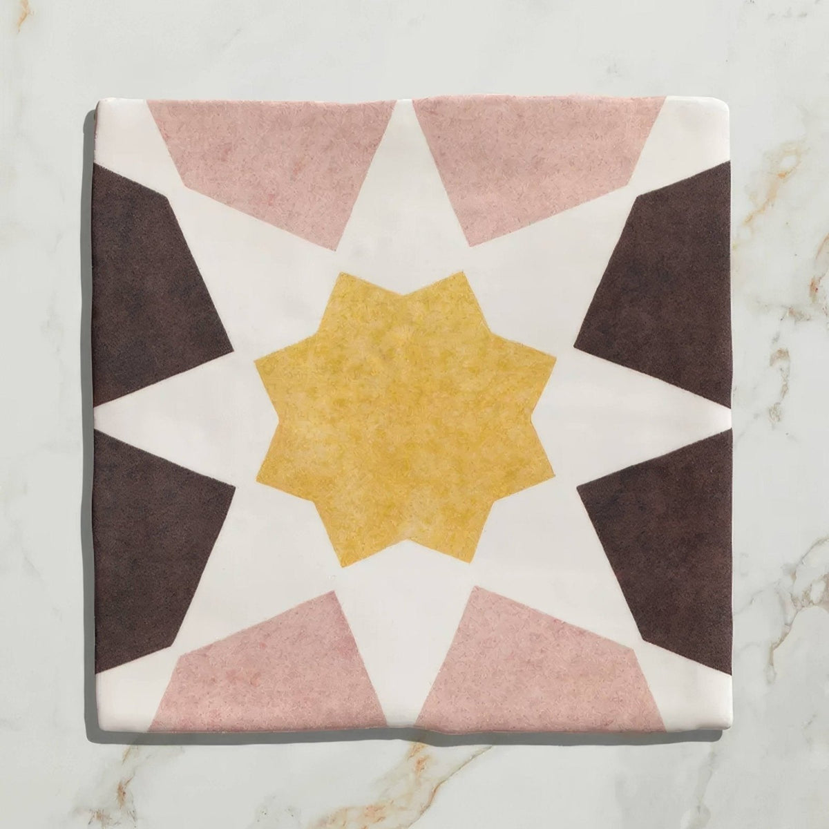 A square ceramic tile with a geometric starburst pattern in muted pink dark brown and mustard yellow tones from Studio Souk
