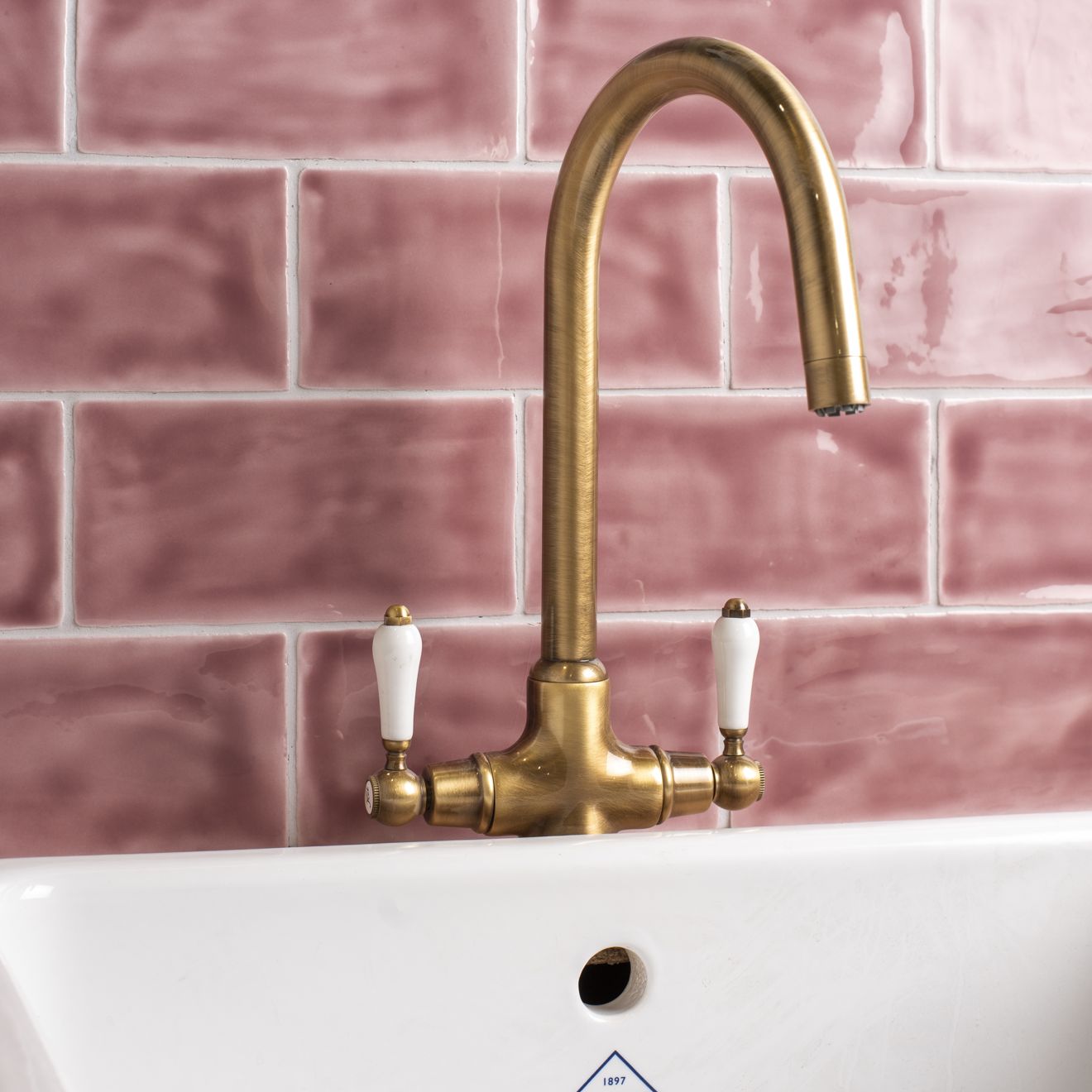 Brass faucet with white ceramic handles set against tayberry brick tile pink tiled backdrop