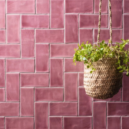 A woven wicker hanging planter in natural beige suspended by braided rope against pink tayberry half tile backdrop