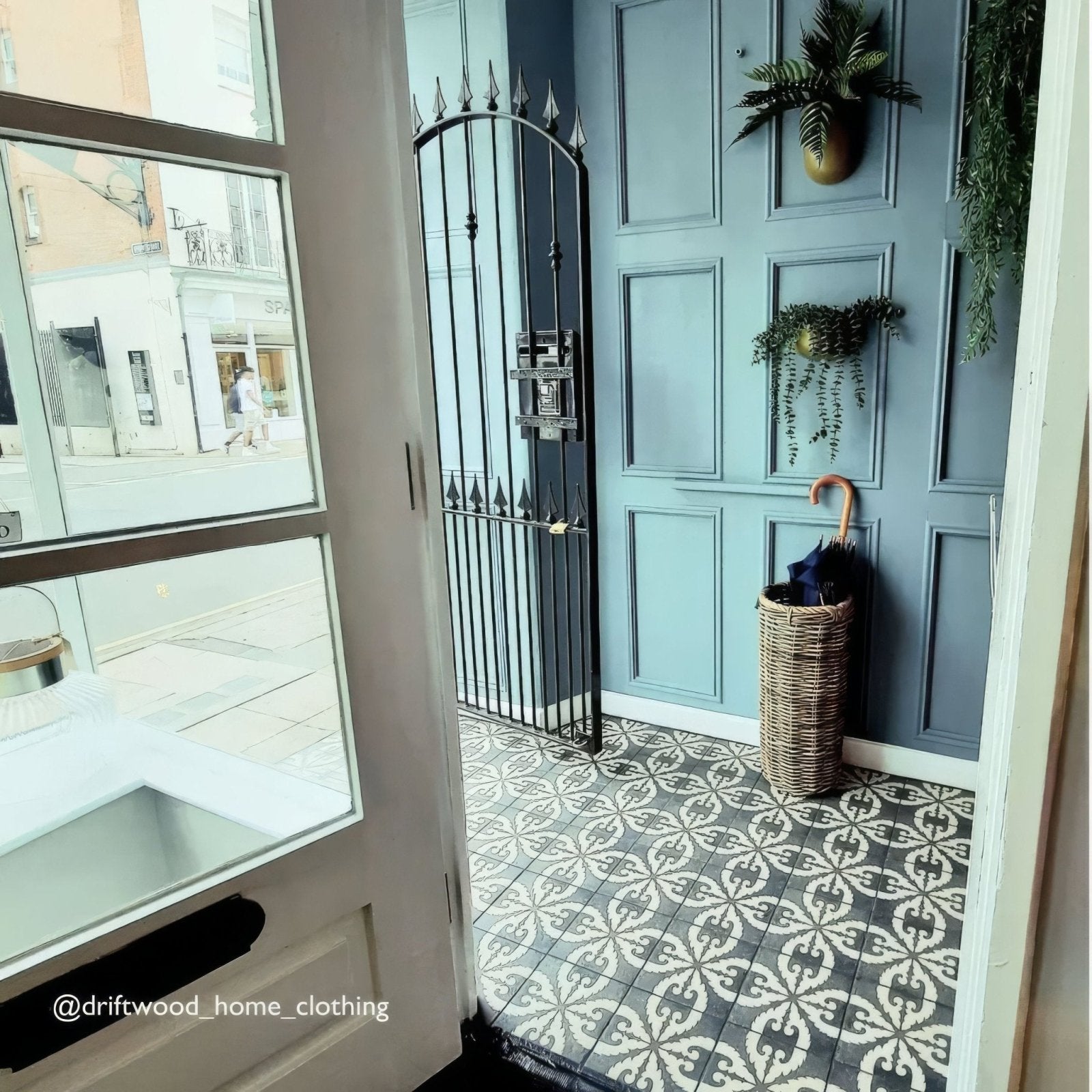 Wicker umbrella stand holding a closed black umbrella with wooden handle on urban velasco patterned porcelain tile