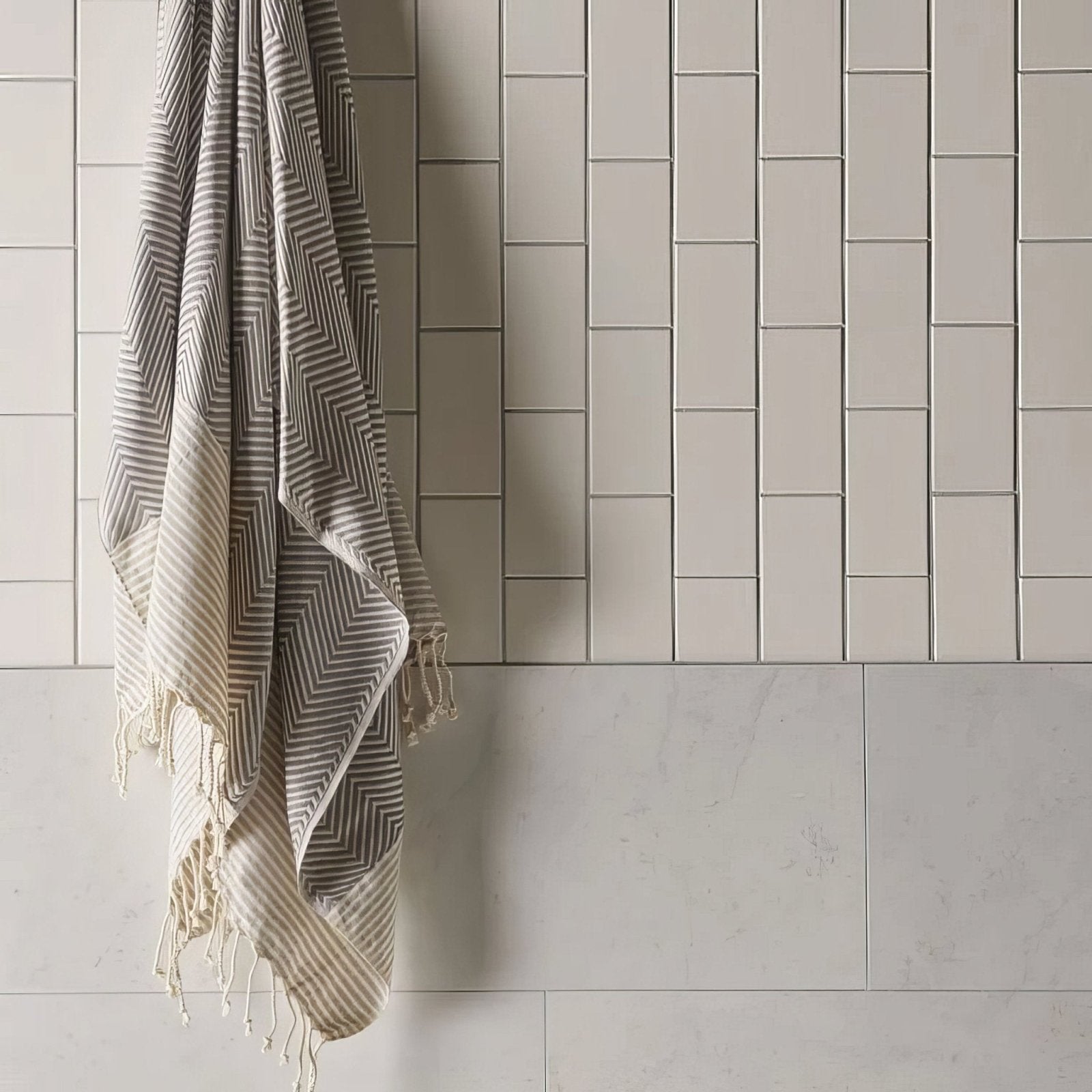 A soft beige and gray geometric patterned vintage white large brick towel with fringed edges hanging on a tiled wall