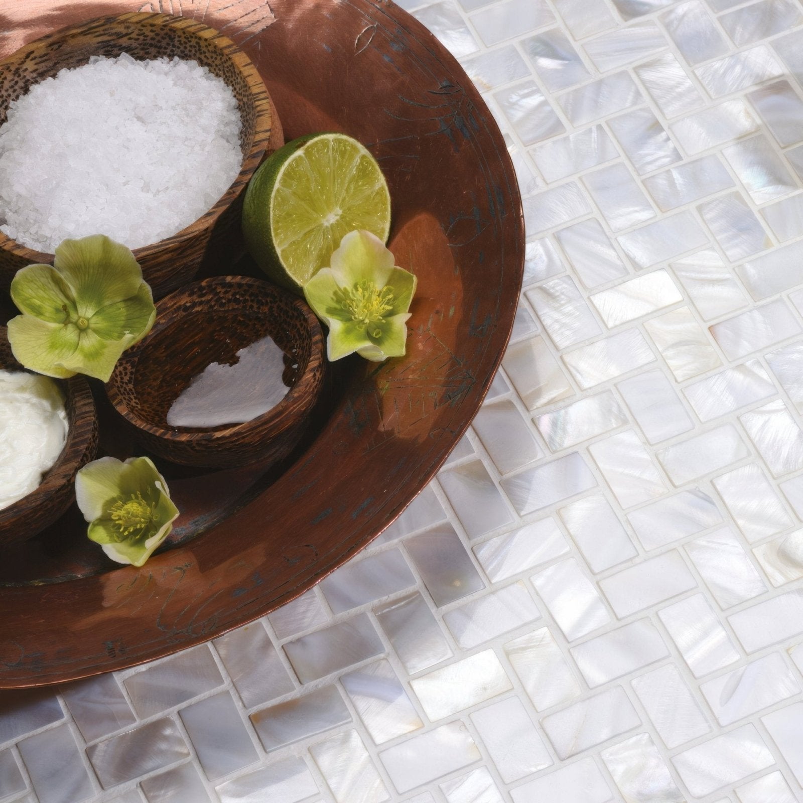 A copper tray with a bowl of coarse white salt, woven bowls of coconut oil and cream, lime slices, and green petals with white pearl herringbone shell mosaic