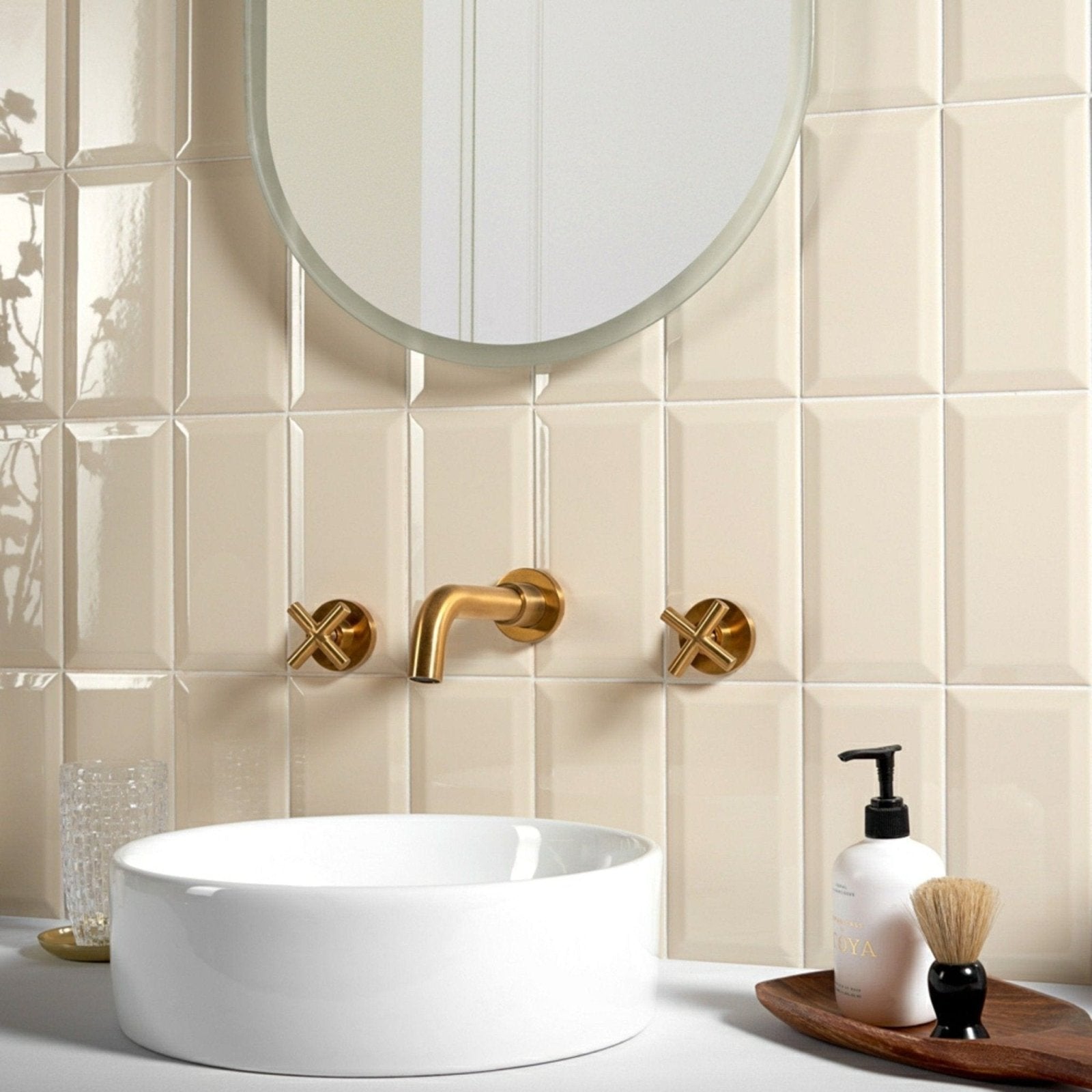 Gold-finished wall-mounted faucet with cross-shaped handles and curved spout set against cream textured Metro Cream Gloss tiles