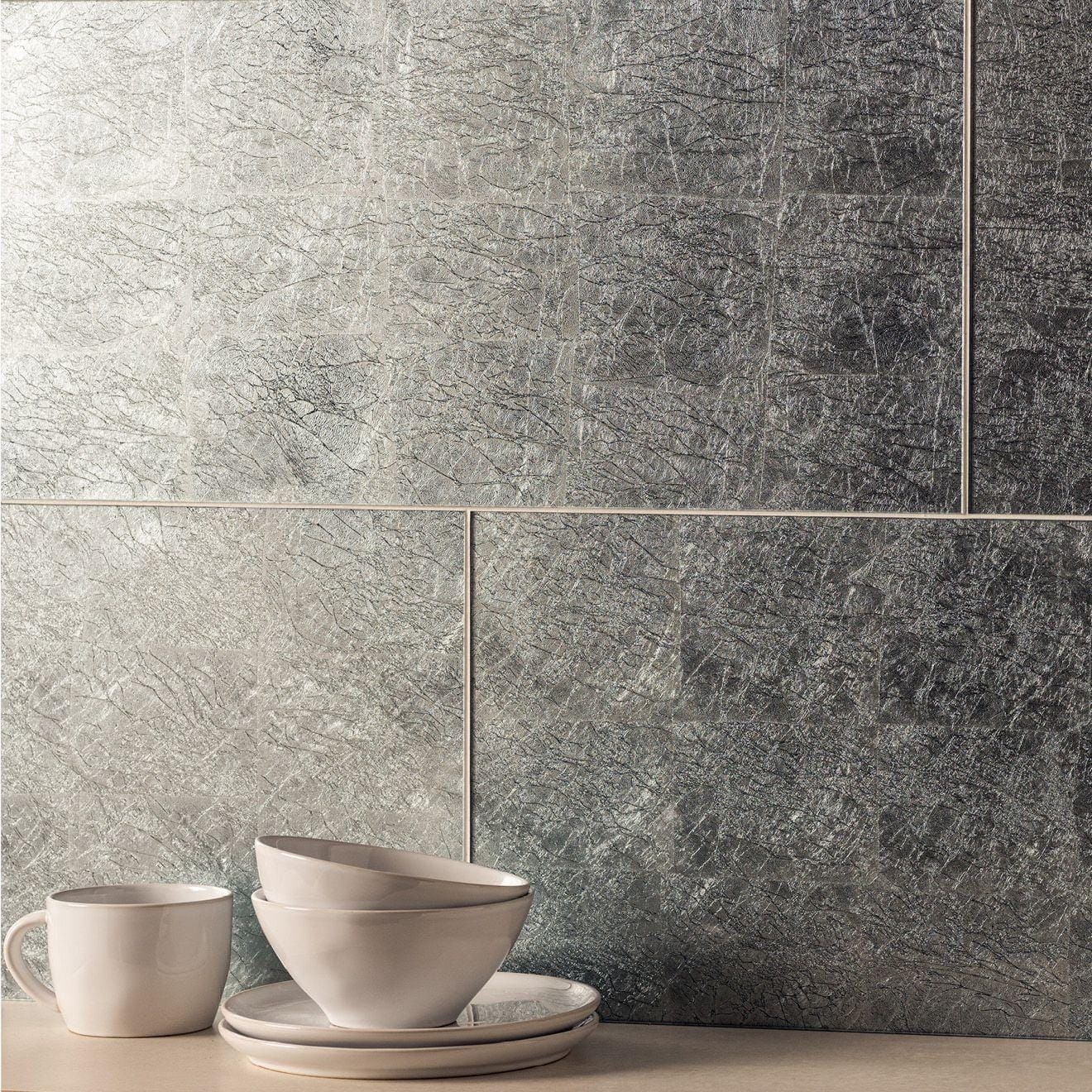 Stacked white ceramic bowls and matching mug rest on plates against silver leaf bold decorative glass tiles backdrop