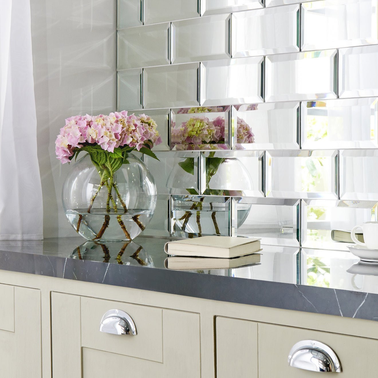 A clear glass vase with pink hydrangeas on a dark countertop next to a silver mirror bevel frame