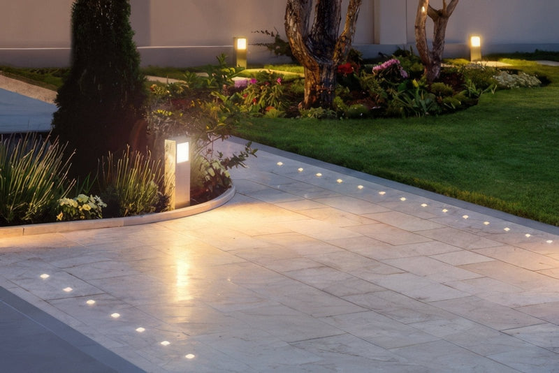 Beige stone outdoor flooring tiles with embedded recessed lights and bordering greenery on a stone pathway.