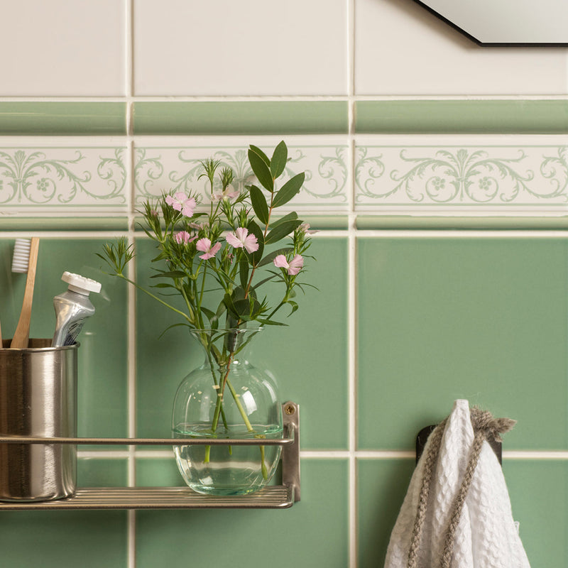 A clear glass vase with pink flowers and green stems on a metal shelf against mint green tiled walls