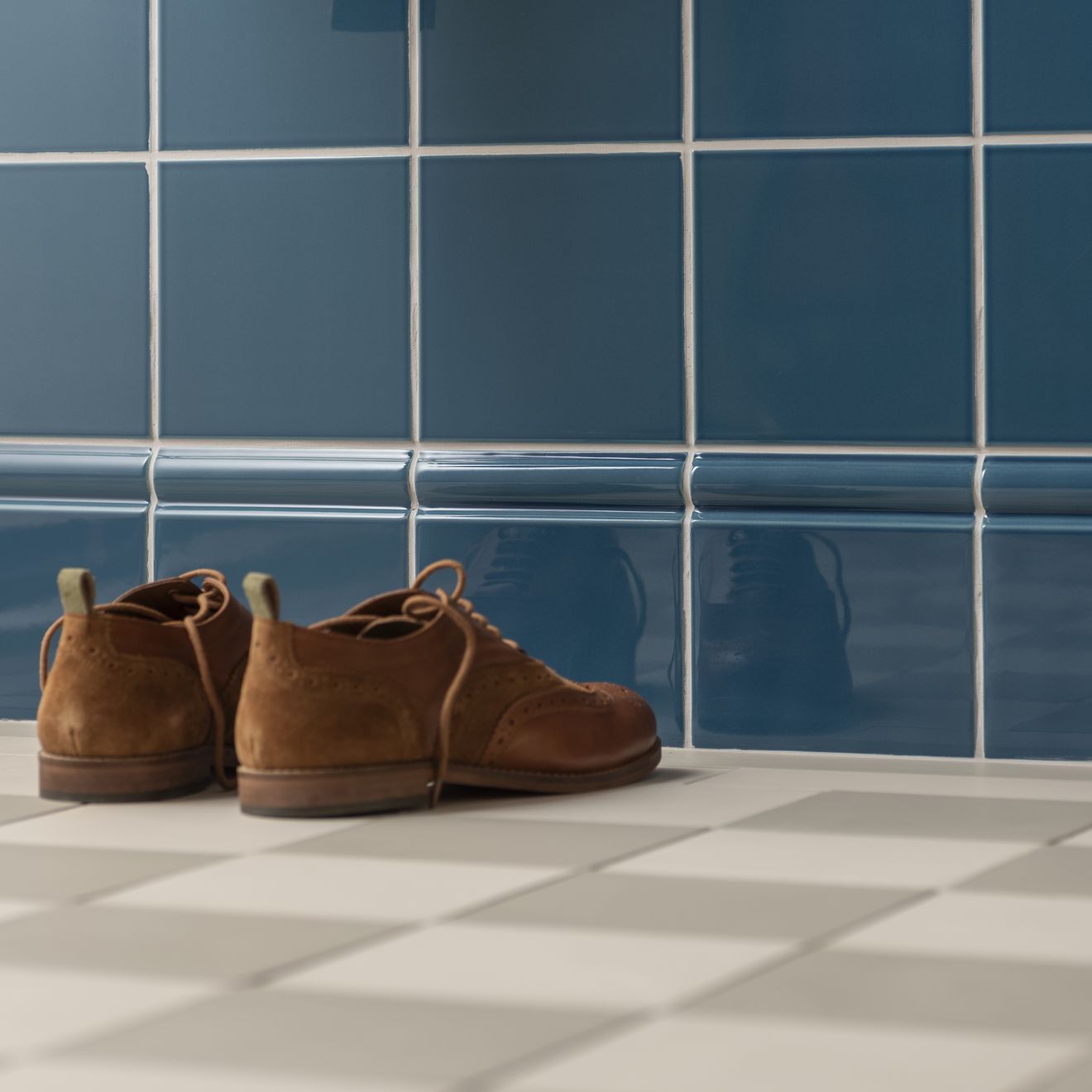 A pair of brown suede lace-up brogue shoes on a checkered floor with blue tiled walls in artworks skirting tile.