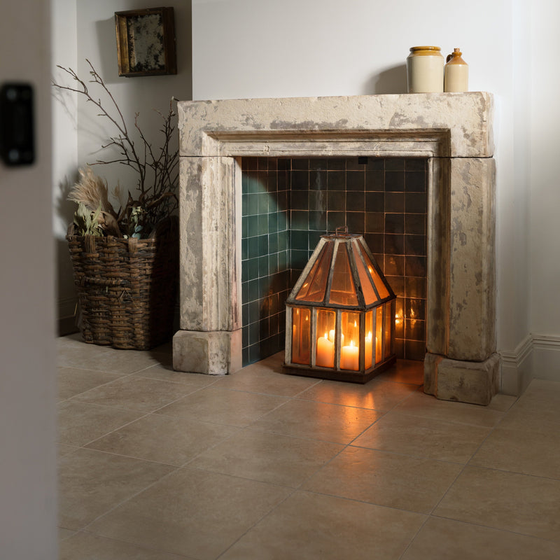 A warm amber-lit geometric glass lantern with a metal frame sits before an antique stone fireplace in ascoli porcelain collection