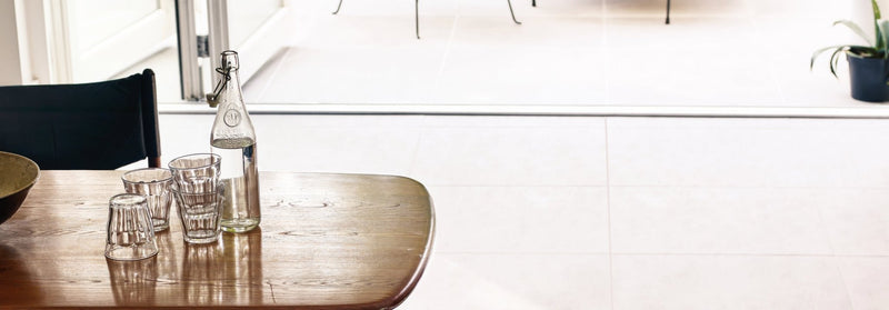 Clear glass water bottle with silver metal stopper and embossed logo standing on wooden table in Barbican Porcelain Tile collection