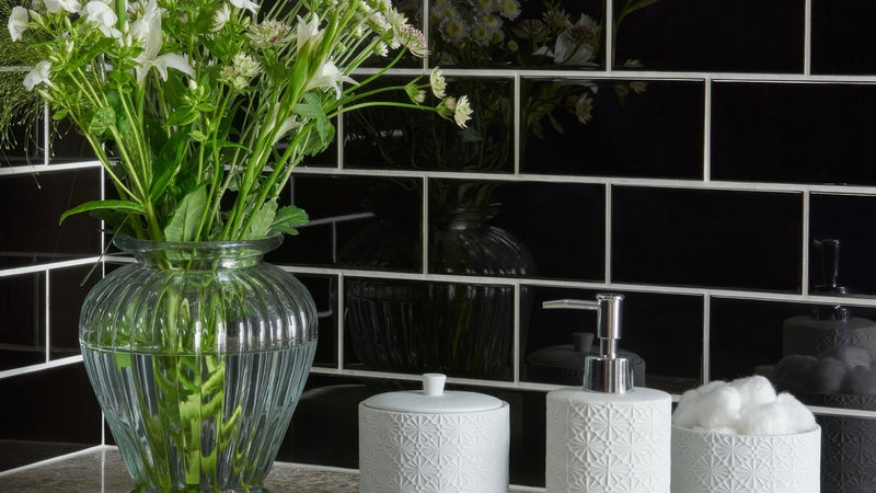 Clear ribbed glass vase holding white flowers displayed on black wall tiles background.
