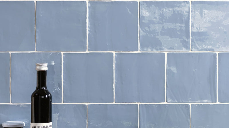 A sleek black glass bottle with silver cap and white label featuring aceto balsamico in blue wall tiles collection