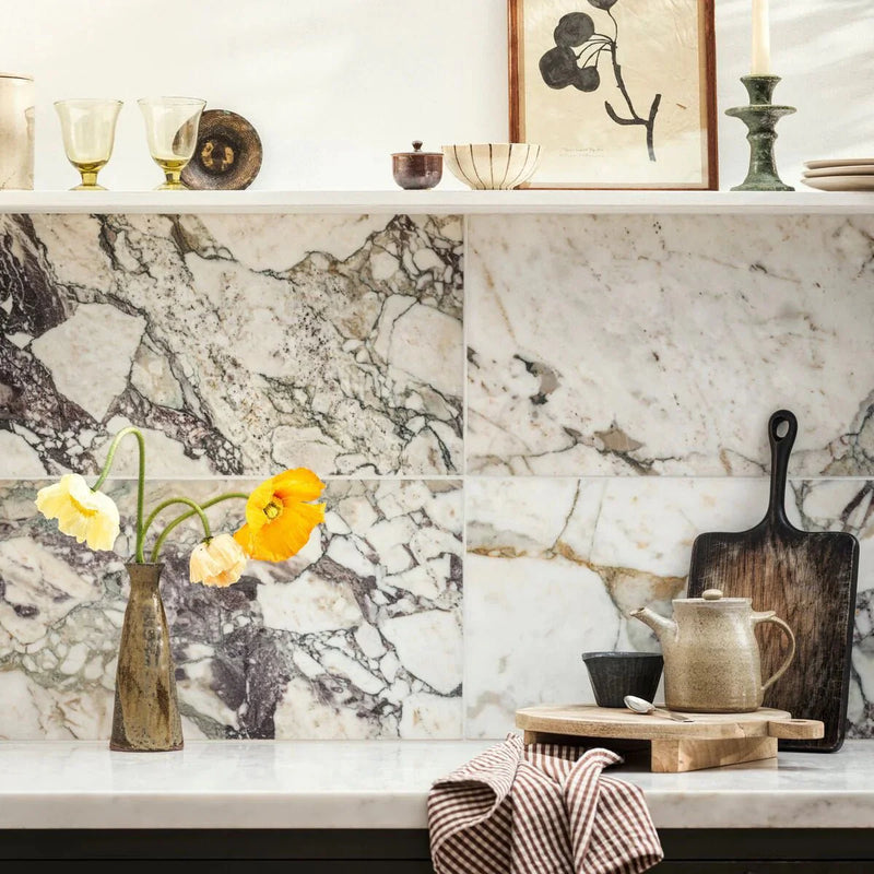 A slender metallic vase with a textured surface holding yellow and orange flowers in calacatta viola honed marble tile setting