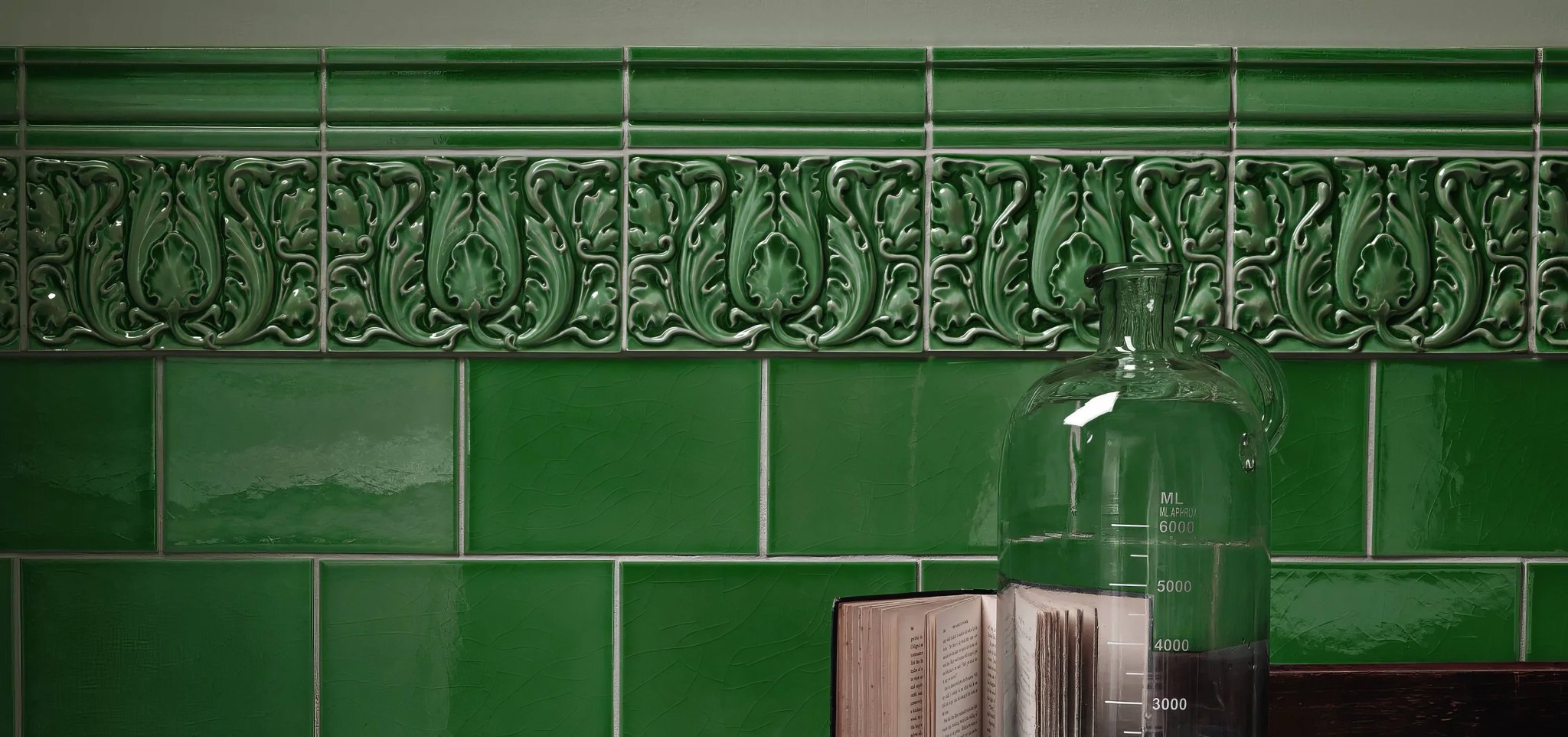 A clear glass measuring jug with graduated markings and narrow neck beside an open book on glossy green Edwardian tiles