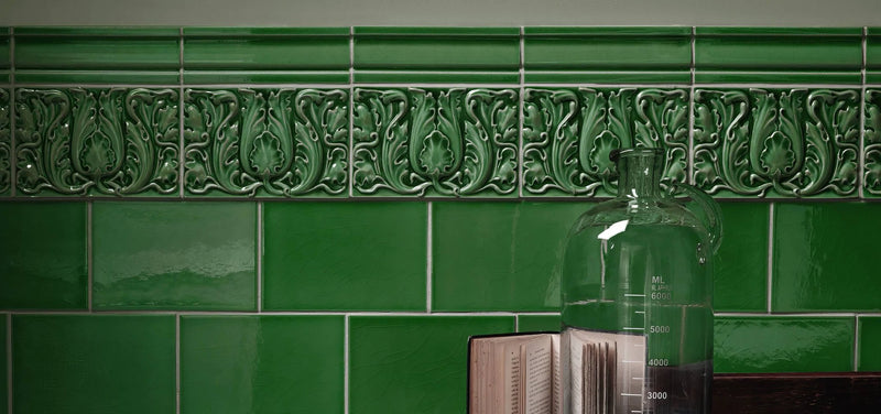 A clear glass measuring jug with graduated markings and narrow neck beside an open book on glossy green Edwardian tiles