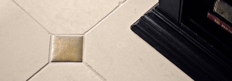 A square beige mosaic tile with a subtle gold tinge at the center placed among larger off-white foundry tiles