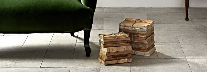 Stacks of aged leather bound books tied with twine on giotto chalk tiles floor beside an emerald green velvet armchair