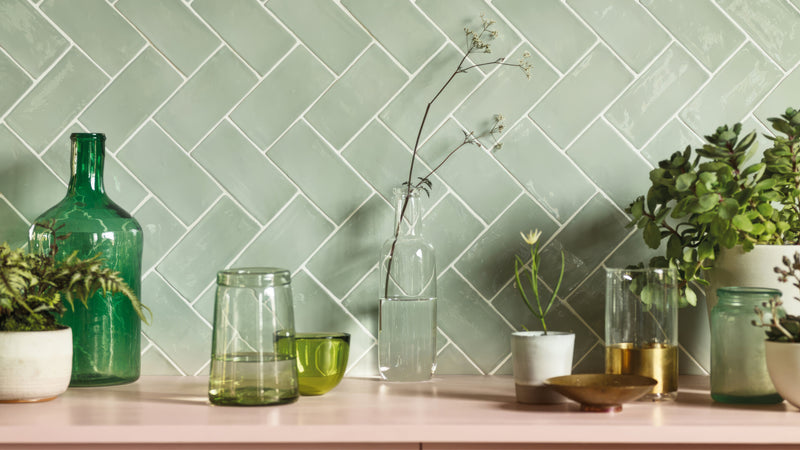 A collection of green and clear glass bottles vases and jars on a pink countertop with herringbone mint green tiles