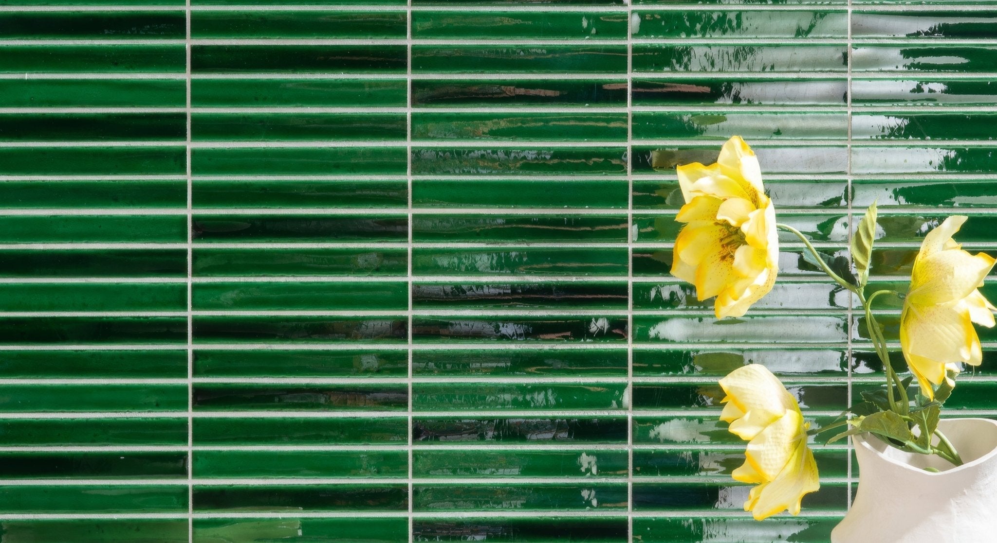 A white ceramic vase holding yellow flowers against glossy green rectangular tiles from the koeda mosaic collection